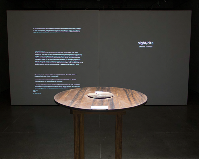 A four legged table with a circular top and circular book stands in front of a wall featuring projected text.