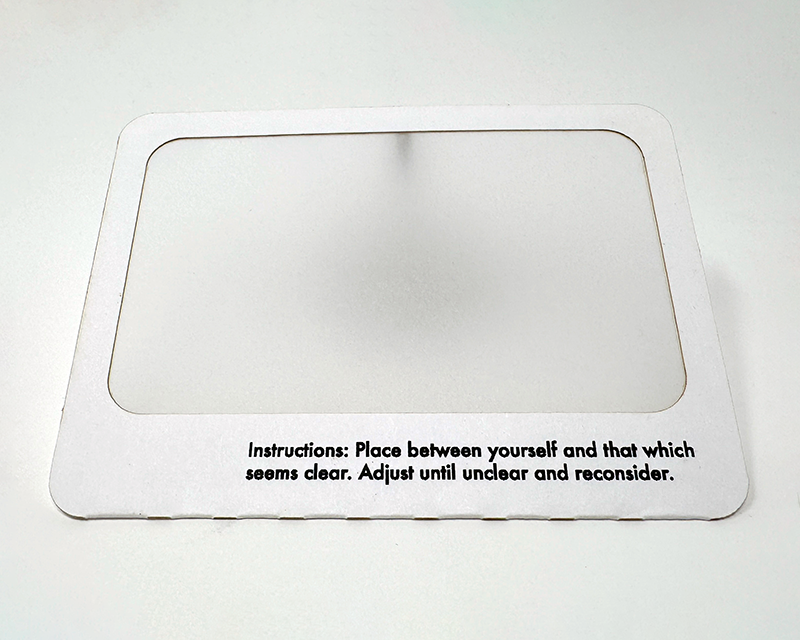 A white paper frame with a translucent paper interior. The bottom of the frame has black text which says 'Instructions: Place between yourself and that which seems clear. Adjust until unclear and reconsider.'